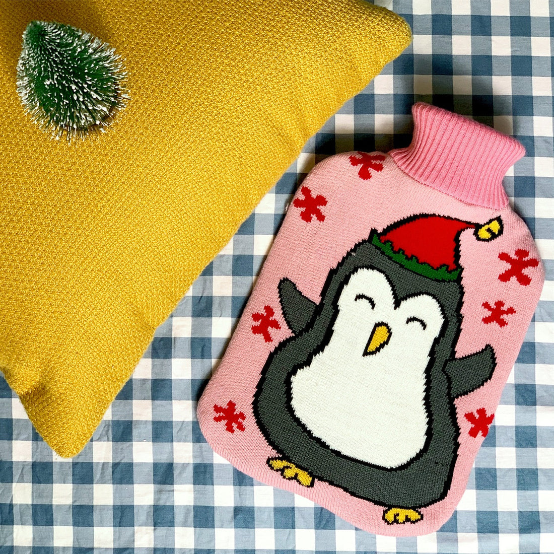 OliviaLiving Christmas Penguin covered hot water bottle on a checked surface beside a yellow cushion